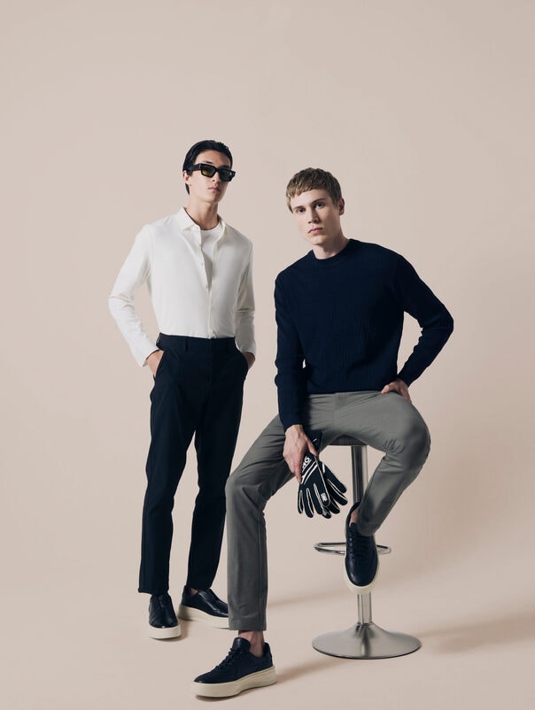 Dois homens de camisa e calça de alfaiataria, um em pé e outro sentado num banco alto