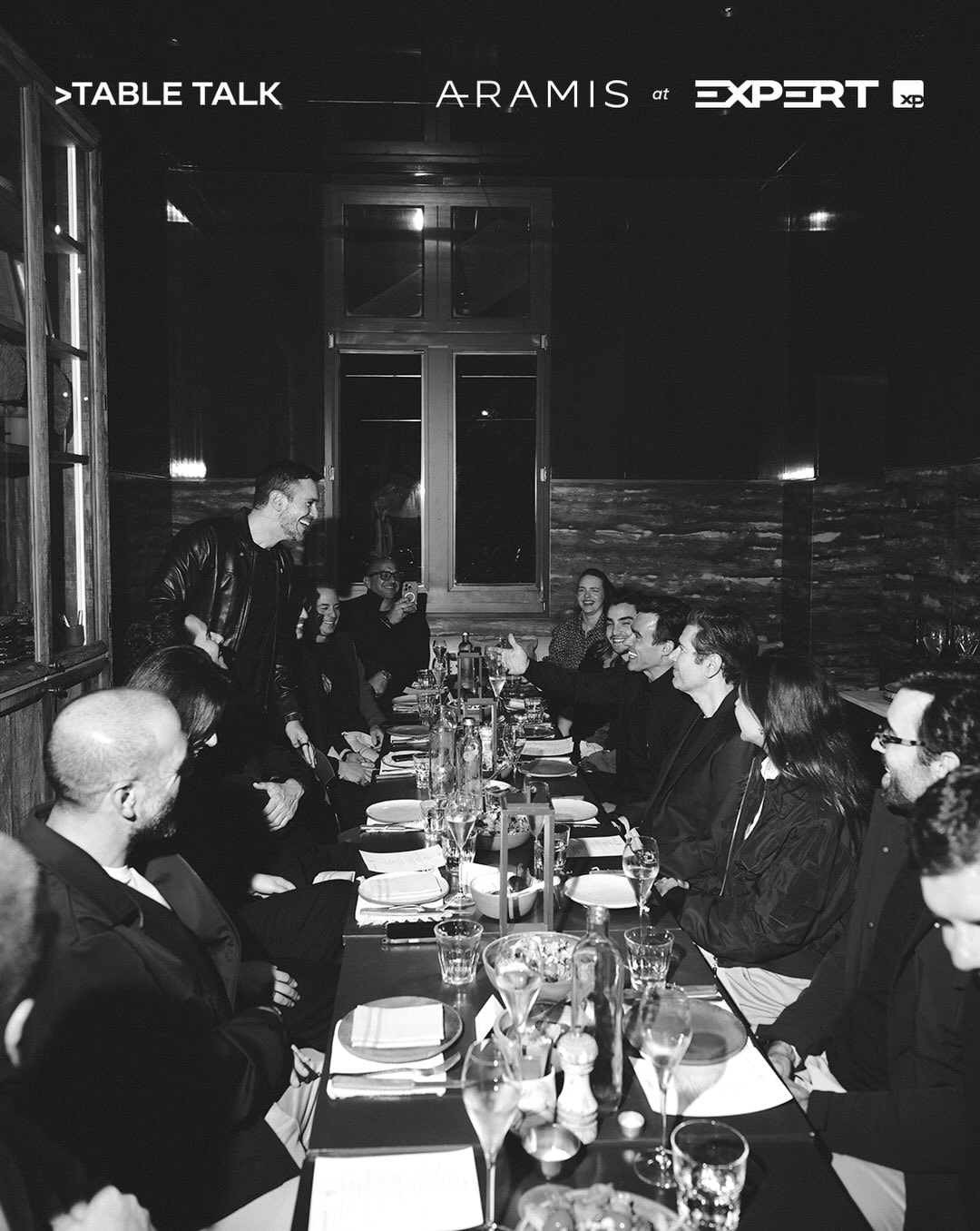 Convidados de um jantar da Aramis reunidos em volta de uma mesa comprida, em preto e branco