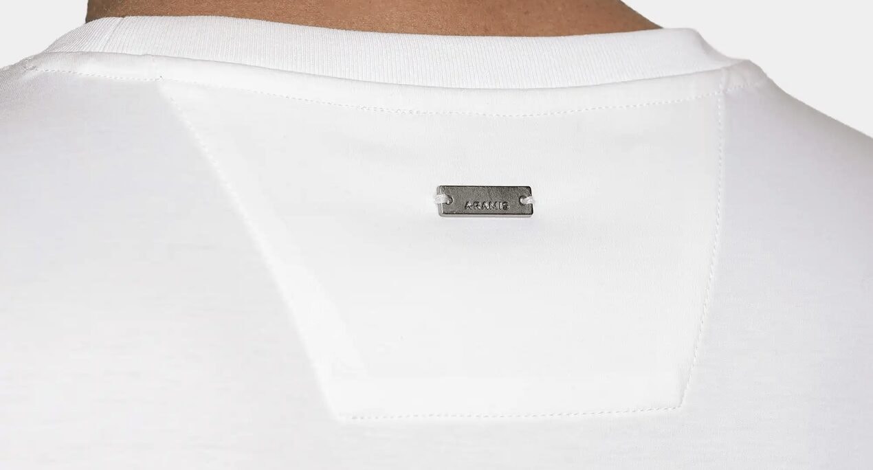 Detalhe das costas de uma camiseta branca, com a etiqueta metálica Aramis costurada logo abaixo da gola