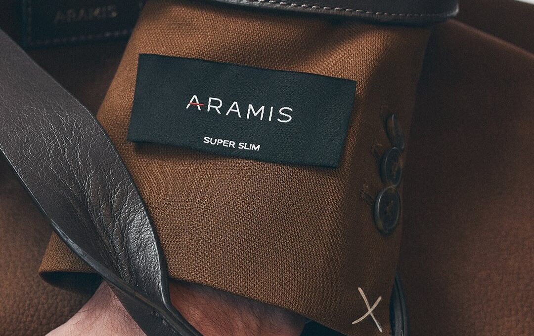 Bolsa de lona marrom com etiqueta Aramis Super Slim, carregada pela alça de couro